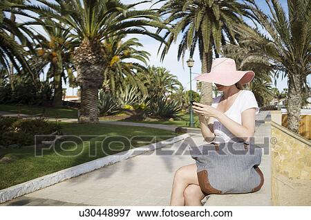 Stock Photo - Mid adult woman texting on smartphone in park gardens at coast. Fotosearch