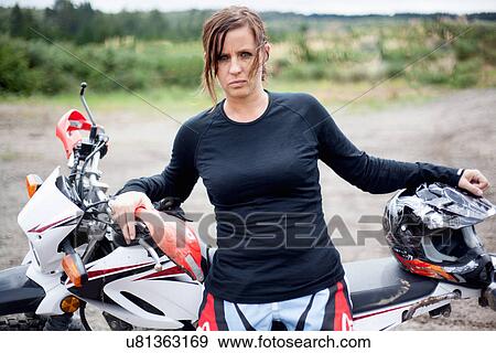 Stock Photo - Portrait of young adult female motorcyclist leaning on motorcycle. Fotosearch