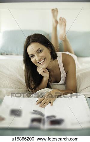 Stock Image - Portrait of young woman lying on bed with magazine. Fotosearch