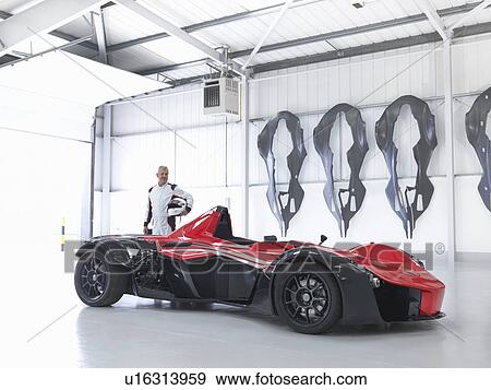Stock Photo - Racing driver with supercar in factory with carbon fibre car body shells on wall. Fotosearch