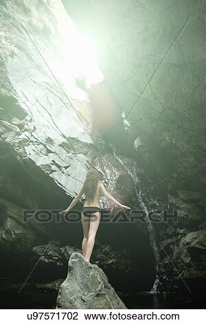 Stock Image - Rear view of young woman standing on rock in river gazing upwards. Fotosearch