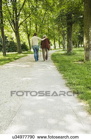 Stock Image - Senior woman and granddaughter walking through park, using walking stick. Fotosearch