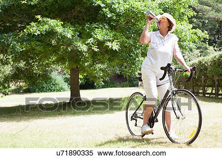 Senior woman cyclist drinking water in park View Large Photo Image Stock Photography - Senior woman cyclist drinking water in park. Fotosearch