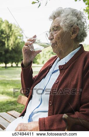 Stock Image - Senior woman sitting on park bench, drinking glass of water. Fotosearch