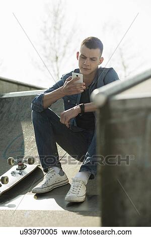 Stock Photography - Skateboarder sitting on ramp, using smartphone. Fotosearch