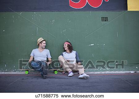 Stock Photography - Two adult male friends sitting on skateboards chatting. Fotosearch