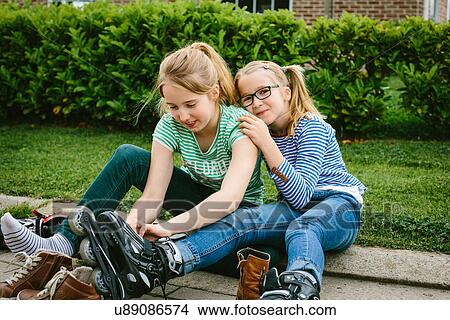 Picture - Two sisters sitting on sidewalk putting on rollerblades. Fotosearch