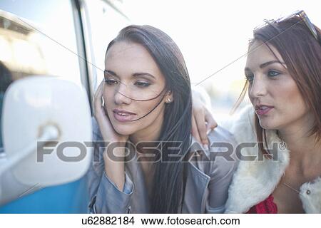 Picture - Two young adult female friends looking in camper van wing mirror. Fotosearch