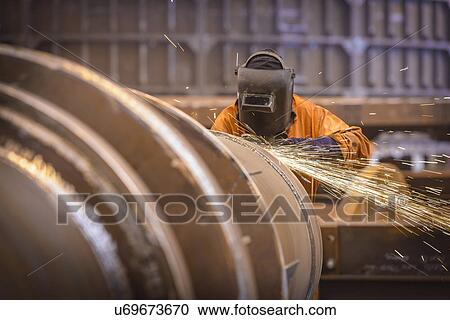Stock Image - Worker grinding metal construction in marine fabrication factory. Fotosearch