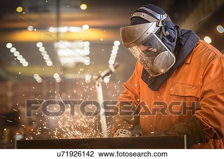 Stock Image - Worker grinding metal construction in marine fabrication factory. Fotosearch