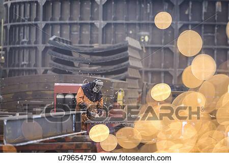 Stock Image - Worker grinding metal construction in marine fabrication factory. Fotosearch