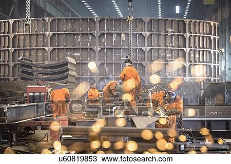 Stock Image - Workers grinding metal construction in marine fabrication factory. Fotosearch