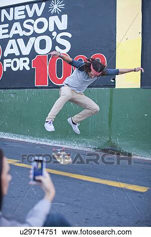 Stock Image - Young adult male being photographed on smartphone doing skateboarding trick. Fotosearch