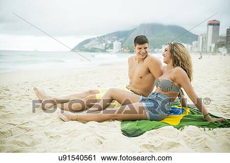 Stock Image - Young couple sitting on brazilian flag, Ipanema Beach, Rio de Janeiro, Brazil. Fotosearch