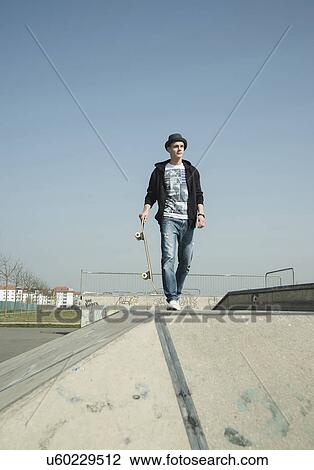 Stock Image - Young man walking through skatepark, carrying skateboard. Fotosearch