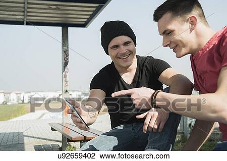 Stock Image - Young men in skatepark, using smartphone. Fotosearch
