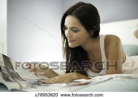 Stock Image - Young woman lying on bed reading newspaper. Fotosearch