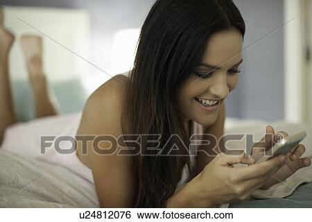 Stock Photograph - Young woman lying on bed texting on smartphone. Fotosearch