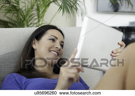 Young woman reclining on sofa using digital tablet View Large Photo Image Stock Image - Young woman reclining on sofa using digital tablet. Fotosearch