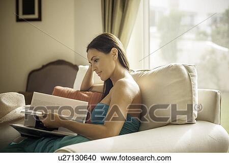 Stock Image - Young woman sitting in armchair reading book. Fotosearch