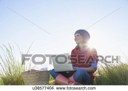 Picture - Young woman sitting in long grass using laptop. Fotosearch
