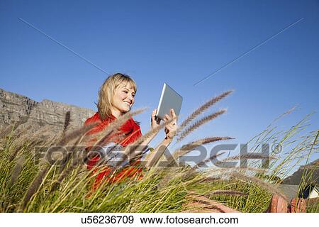 Young woman sitting in long grass using digital tablet View Large Photo Image Stock Photo - Young woman sitting in long grass using digital tablet. Fotosearch