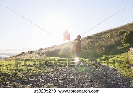 Stock Image - Young woman strolling along dirt track with bunch of balloons. Fotosearch