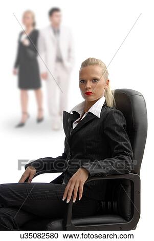 Stock Image - Business woman sitting in a chair with co-workers in background. Fotosearch