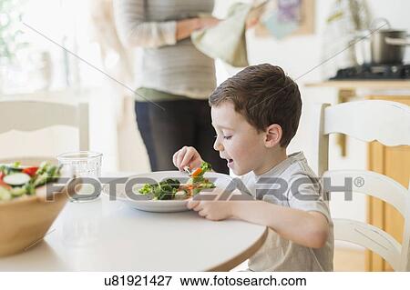 Stock Photo - boy (6-7) eating healthy dinner, mother in background. Fotosearch