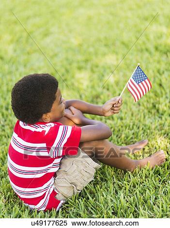 Stock Photography - boy (6-7) sitting on grass and holding american flag. Fotosearch