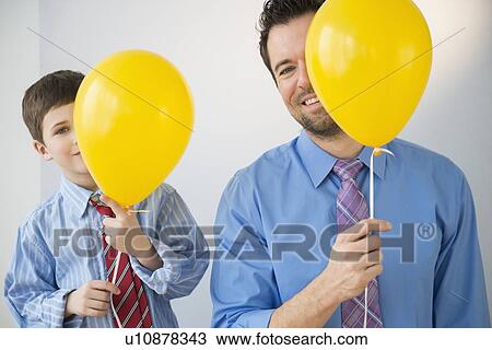 Stock Image - Father and son (8-9) peeking behind yellow balloons. Fotosearch