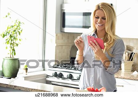 Stock Photo - Happy woman text messaging in kitchen. Fotosearch