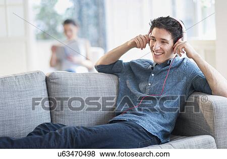 Stock Photo - Man listening to music on sofa, woman in background. Fotosearch