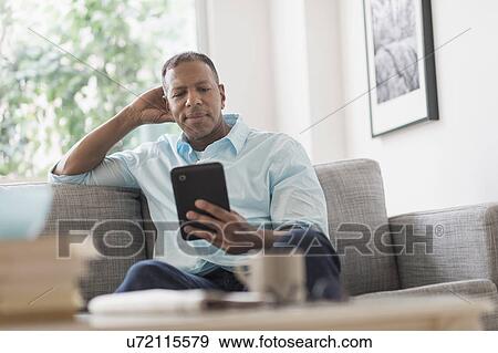 Stock Photo - Man sitting on sofa at home and using digital tablet. Fotosearch