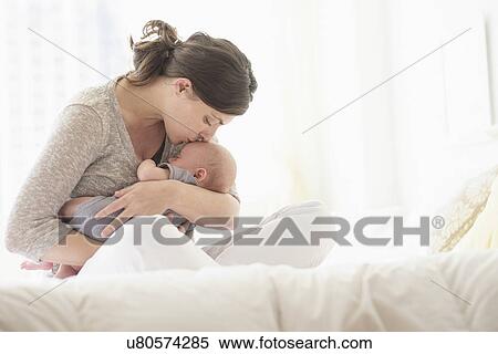 Stock Photography - Mother kissing baby boy (2-5 months) in bed. Fotosearch