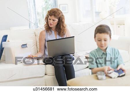 Stock Image - Mother using laptop, son (6-7) playing with toy cars. Fotosearch