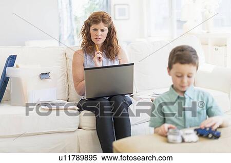 Stock Photography - Mother using laptop, son (6-7) playing with toy cars. Fotosearch