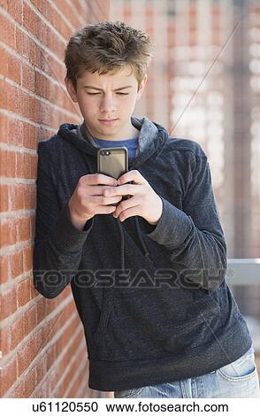 Stock Image - Teenage boy (16-17) leaning against brick wall texting. Fotosearch