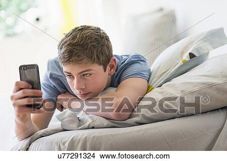 Picture - Teenage boy (16-17) lying on bed and text messaging. Fotosearch