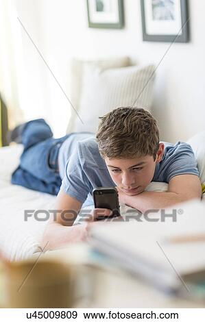 Stock Photo - Teenage boy (16-17) lying on bed and text messaging. Fotosearch