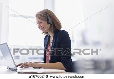 Stock Photograph - Young office worker talking on headset and typing. Fotosearch
