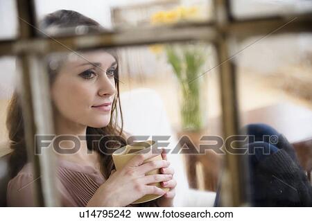 Stock Image - Young woman contemplating in living room with coffee cup in hands. Fotosearch