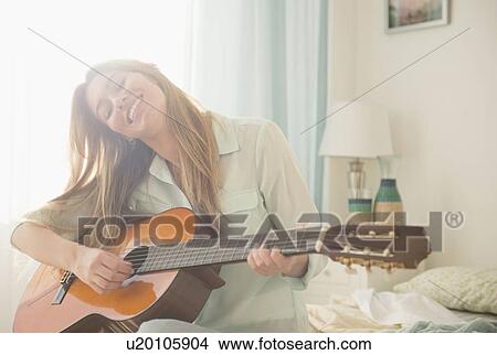 Picture - Young woman playing acustic guitar on bed. Fotosearch
