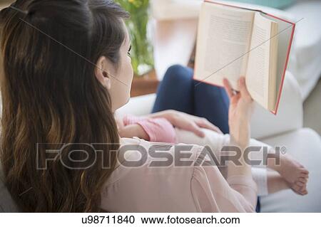 Stock Image - Young woman reading book and holding daughter(6-11 months). Fotosearch