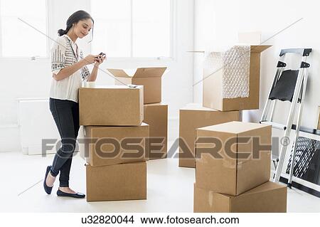 Young woman standing among boxes in new home and using mobile phone View Large Photo Image Picture - Young woman standing among boxes in new home and using mobile phone. Fotosearch