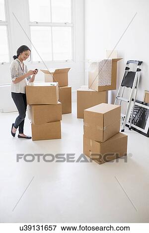 Stock Photo - Young woman standing among boxes in new home and using mobile phone. Fotosearch