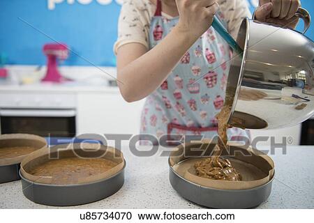 Stock Image - Female hands pouring mixture into cake tins in bakery. Fotosearch