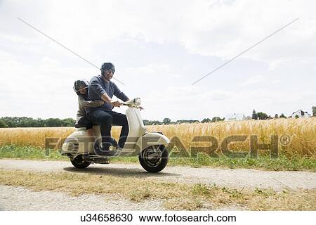 Stock Image - Mature man and daughter riding motor scooter along dirt track. Fotosearch