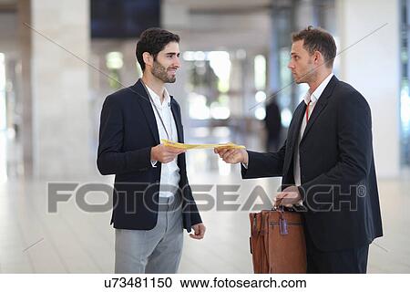 Stock Image - Office worker handing paperwork to businessman in conference centre. Fotosearch