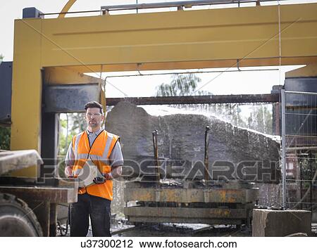 Stock Image - Portrait of quarry worker in front of large rock being cut in stone saw. Fotosearch